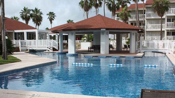 A pool area with lounge chairs, a covered seating area, and a building with balconies.