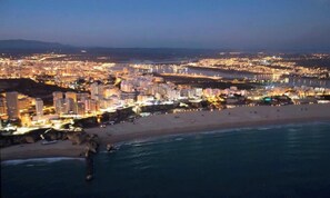 A coastal cityscape at dusk with illuminated buildings, a beach, and a body of water.