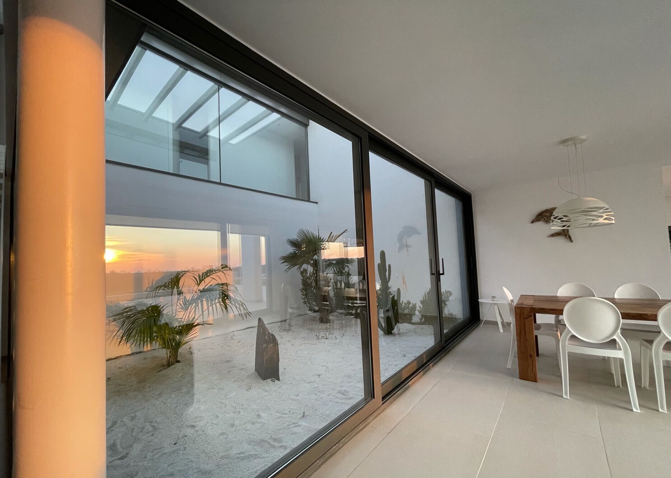 Un intérieur moderne avec un grand mur de verre, un coin repas avec des chaises blanches et un jardin de cactus.