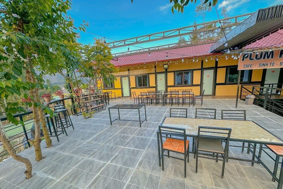 An outdoor café area with tables and chairs, surrounded by trees and a building with a red roof.