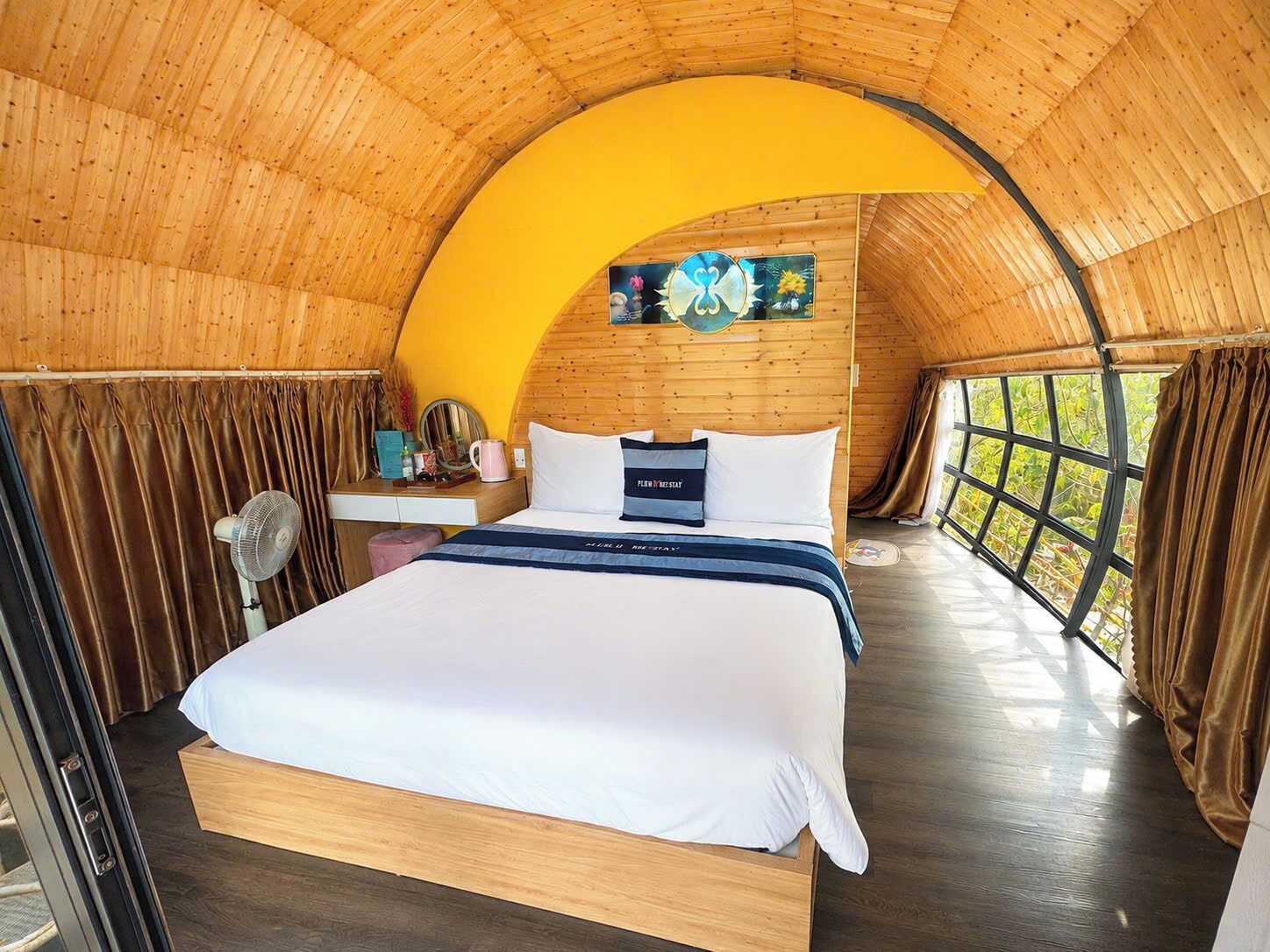 A cozy, circular room with a wooden ceiling and walls, a bed with a white mattress and navy blue bedding, a bedside table with a lamp, and a window with curtains.