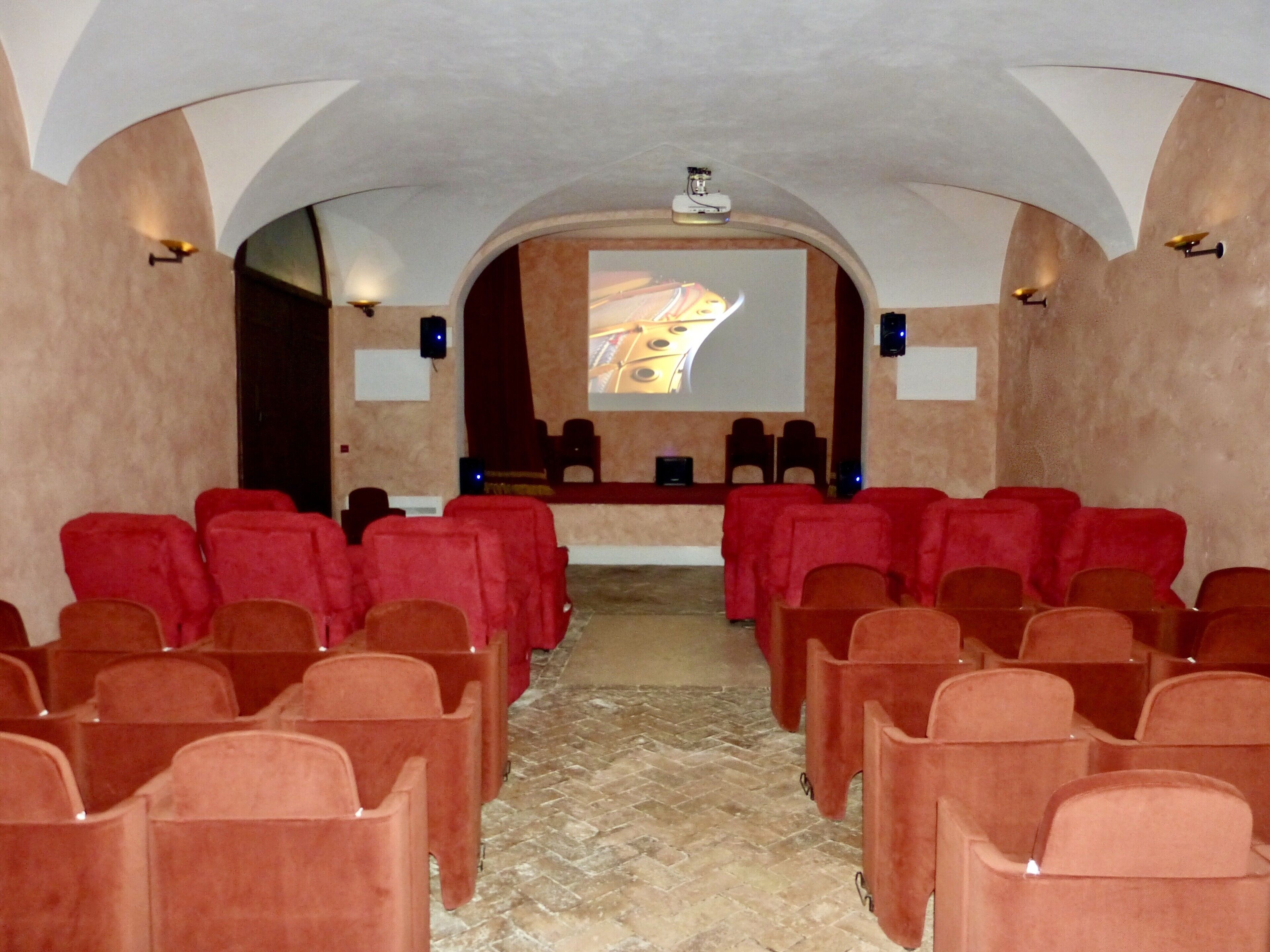 A theater with rows of red and brown chairs, a stage with a projection screen, and a stone floor.