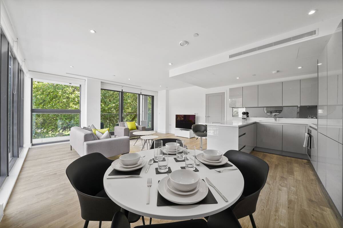 Une cuisine moderne avec des meubles blancs, un coin repas doté d’une table ronde pour quatre personnes, et un salon avec un canapé et un téléviseur.