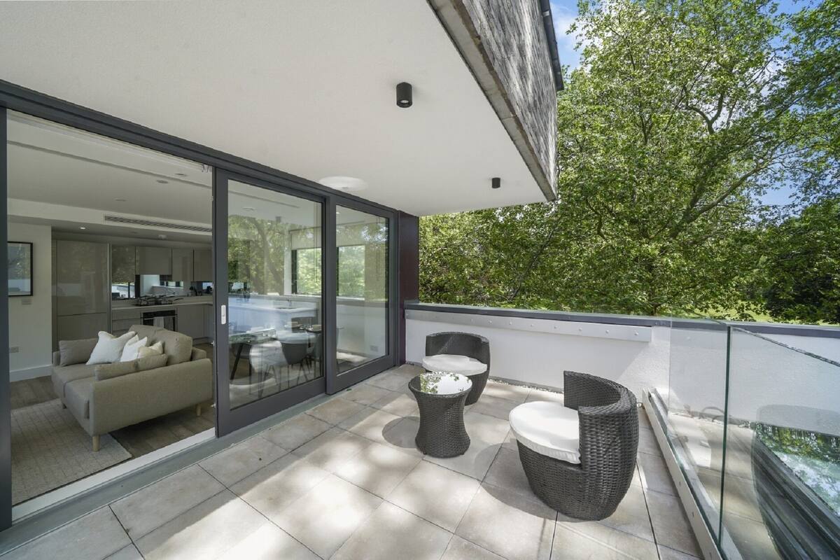 Un balcon moderne avec une porte vitrée donnant sur un espace de vie, des meubles en osier et une vue sur les arbres.