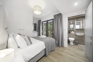 Ein modernes Schlafzimmer mit einem großen Bett, einem Nachttisch, einem Fenster mit Vorhängen, einem Badezimmer mit Waschbecken und Toilette sowie Blick auf Grünflächen.