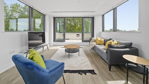 A modern living room with a sofa, a blue armchair, a coffee table, and large windows offering a view of trees and buildings. - Executive Apartments in Bermondsey FREE WIFI & AIRCON by City Stay Aparts London (London)