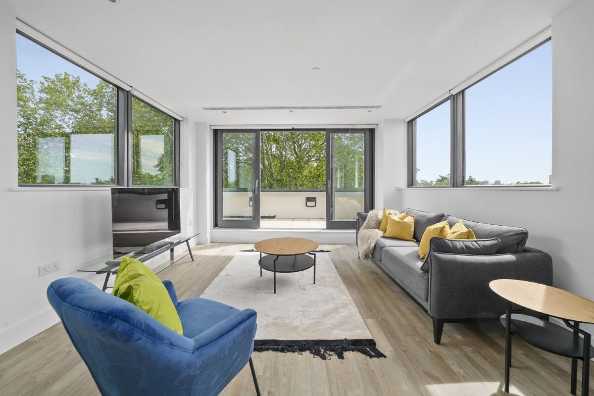 Un salon moderne comprenant un canapé, un fauteuil bleu, une table basse et de grandes fenêtres donnant sur des arbres et des immeubles.