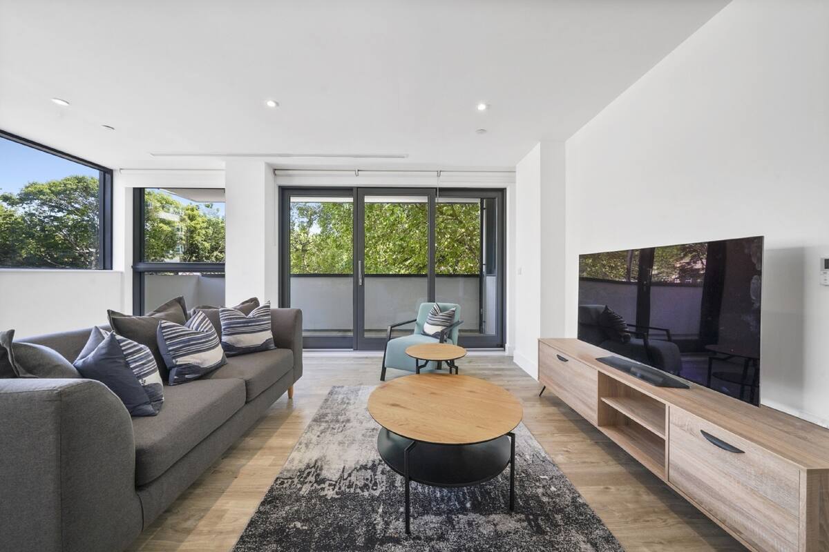 Un salon moderne avec un canapé gris, une table basse ronde, un téléviseur et une grande fenêtre donnant sur des arbres.