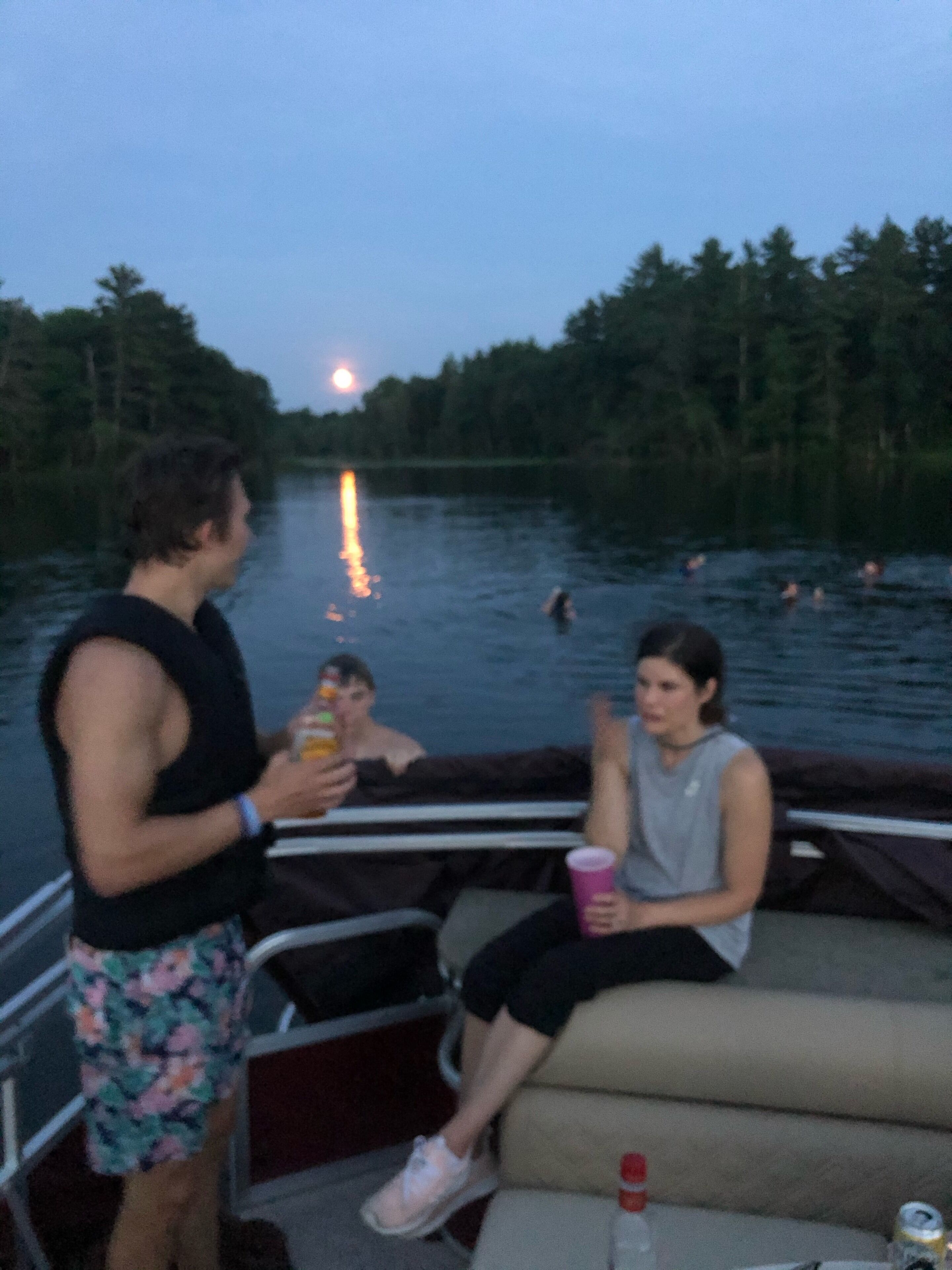A boat with a person standing and two people sitting on the deck, overlooking a body of water with people swimming.