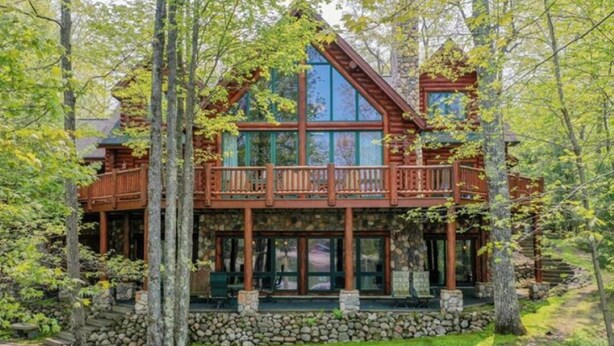 A log cabin with a wooden deck and stone foundation, surrounded by trees. - Luxury 7,500‑Sq‑Ft Lakefront Lodge, Private Dock & Snowmobile Access (Hayward)