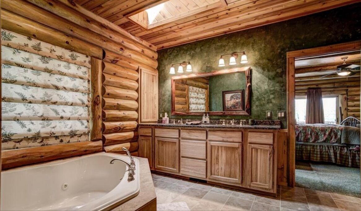 A log cabin bathroom with a large bathtub, wooden cabinets, and a mirror with lights above the sink.