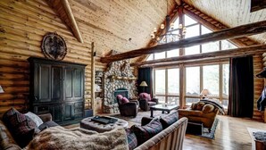 A log cabin interior with a stone fireplace, wooden furniture, and large windows. - Luxury 7,500‑Sq‑Ft Lakefront Lodge, Private Dock & Snowmobile Access (Hayward)