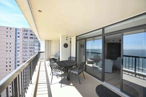 Un balcón con una mesa y sillas, una puerta de vidrio que da a una habitación con vista al mar, y una barandilla.