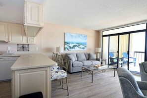 A modern living room with a sofa, coffee table, and chairs. There is a kitchen area with cabinets and a dishwasher. A large window provides a view of the outdoors.