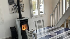 Un comedor con una mesa cubierta por un mantel a rayas, sillas blancas, un calentador negro, una ventana con vistas al exterior y una bicicleta colgada en la pared.