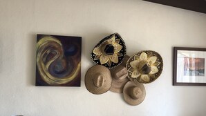 A living room with a beige sofa, patterned cushions, a glass table with a lamp, and decorative wall art including hats and a swirl painting. - LaBamba - Great Condo for Adventurer in beautiful San Jose del Cabo (San José del Cabo)