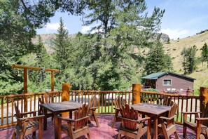 A wooden deck with tables and chairs, overlooking a forested hillside and a cabin.