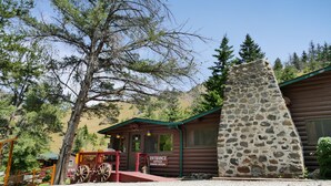 A rustic log cabin with a stone chimney, a wooden porch, and a sign indicating an entrance office and dining room.