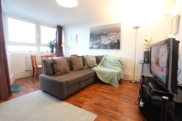 A modern living room with a grey sofa, wooden floor, a large window, and a television. - Bright Comfortable Chelsea flat - Great Location! (London)