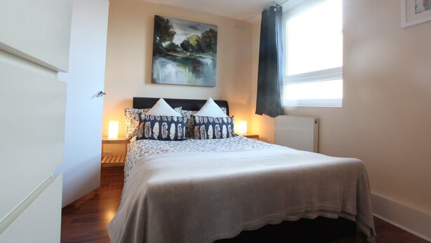Une chambre à coucher avec un lit, un tableau au mur, une fenêtre avec des rideaux et un radiateur.