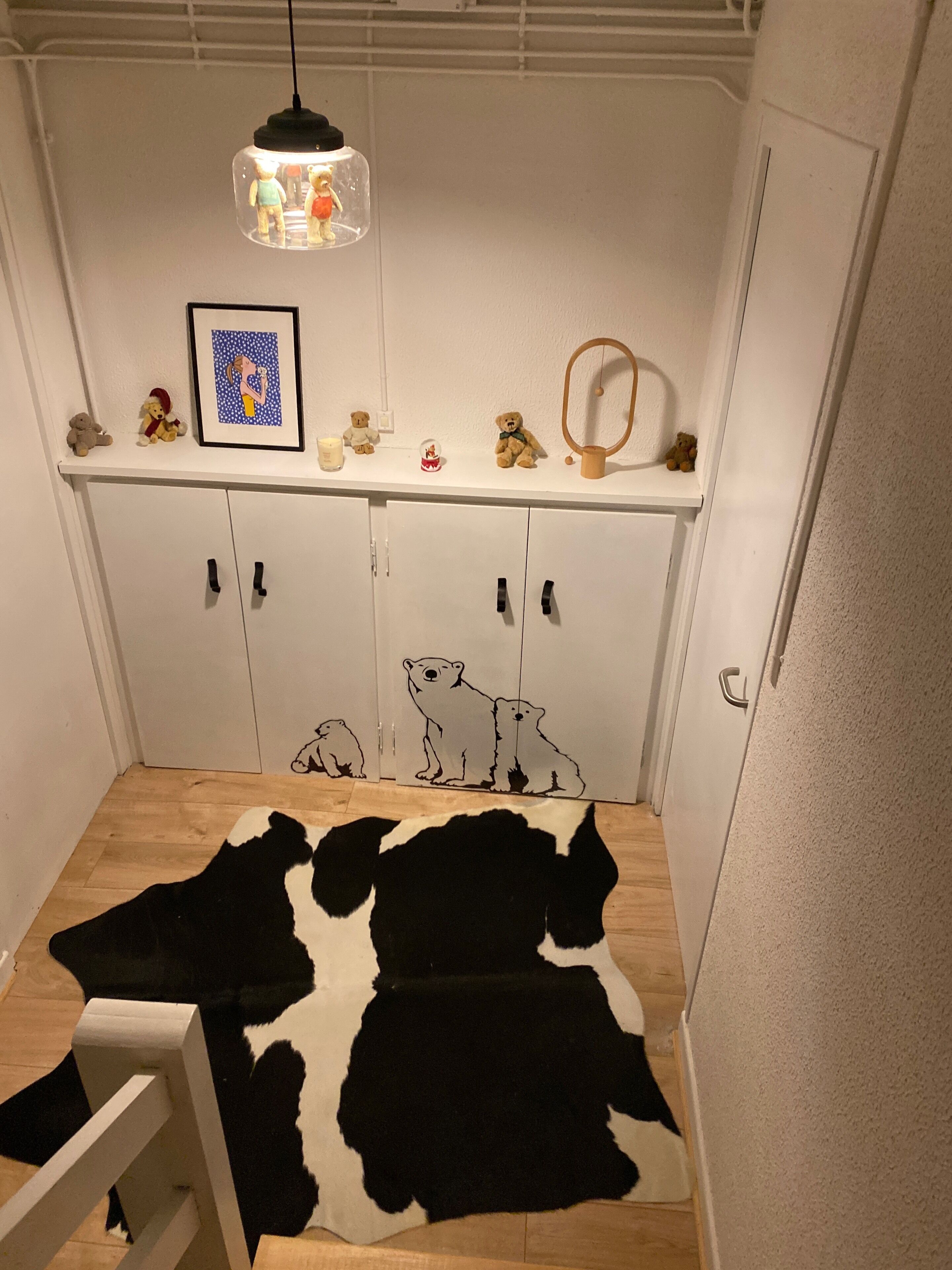 Une pièce avec une armoire blanche, une petite table surmontée d’un tapis en peau de vache et une étagère où sont disposés divers objets, dont un ours en peluche et un tableau encadré.