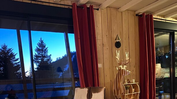 Ett mysigt rum med ett stort fönster, en trästol med en fäll över, ett litet bord med ljus och en röd gardin.