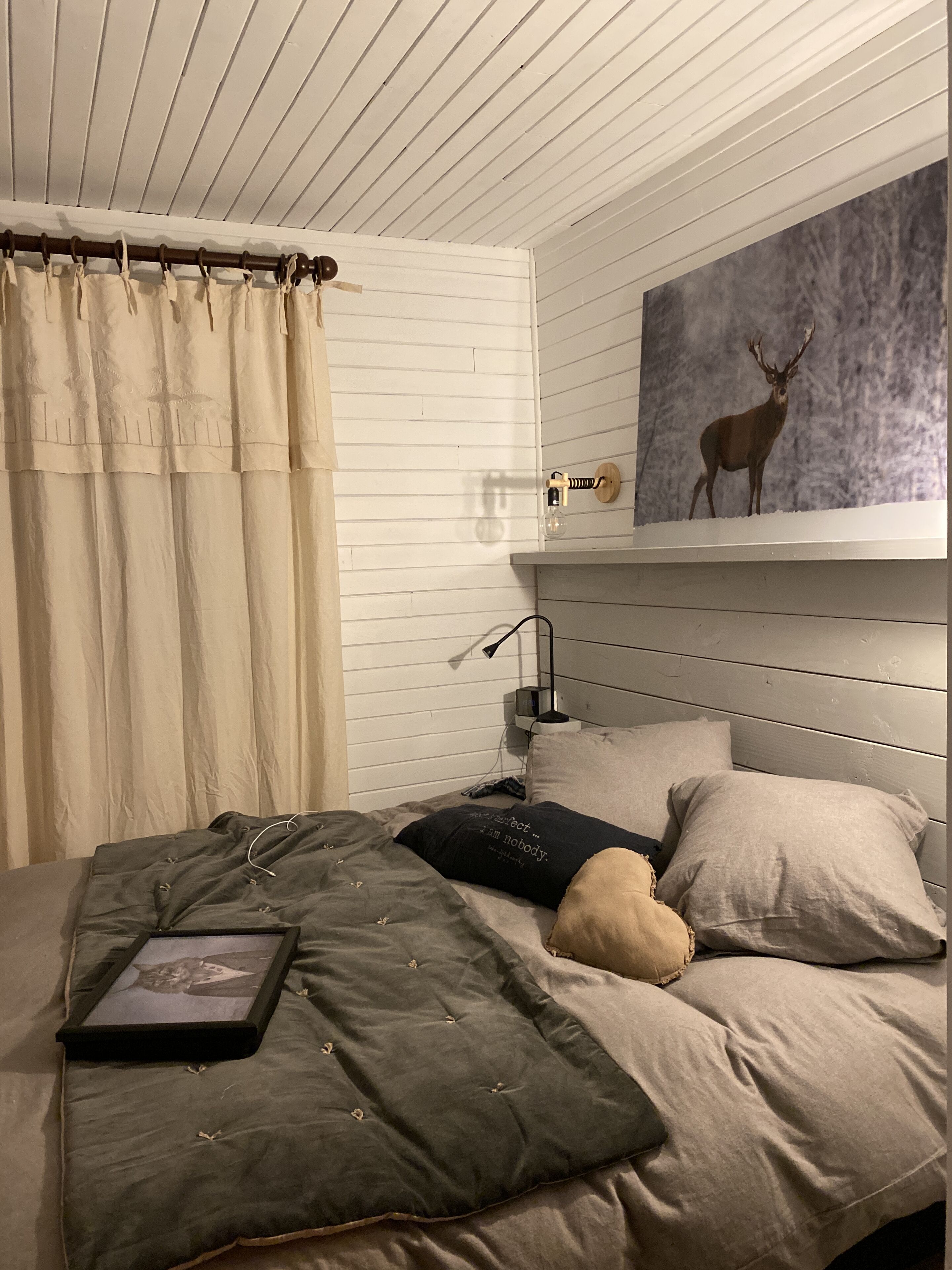 A bedroom with a bed, a framed picture of a cat, a lamp, and a wall-mounted shelf with a deer painting.