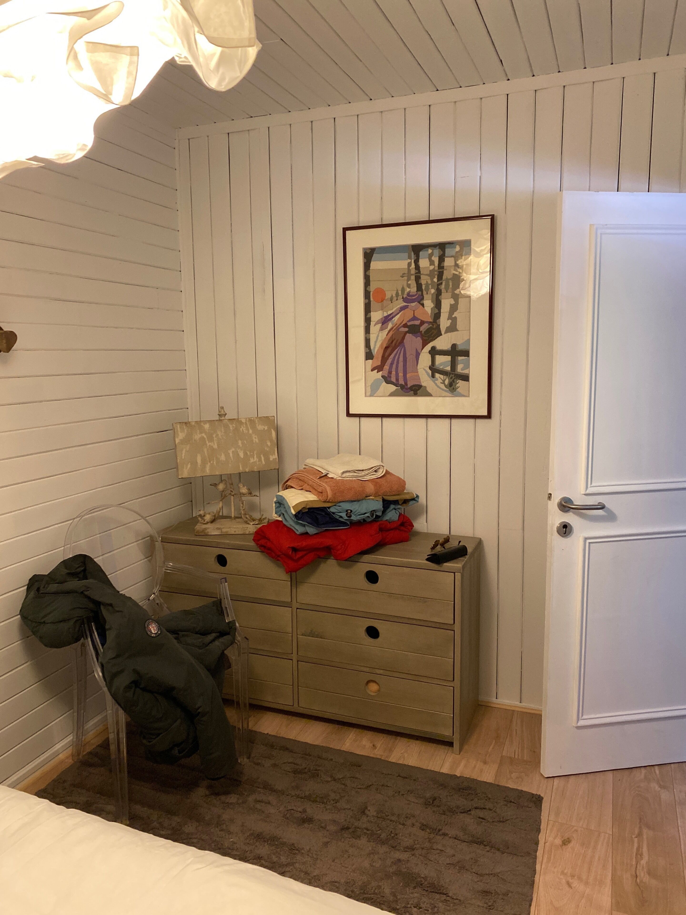A room with a wooden dresser, a framed picture on the wall, a lamp, and a door.