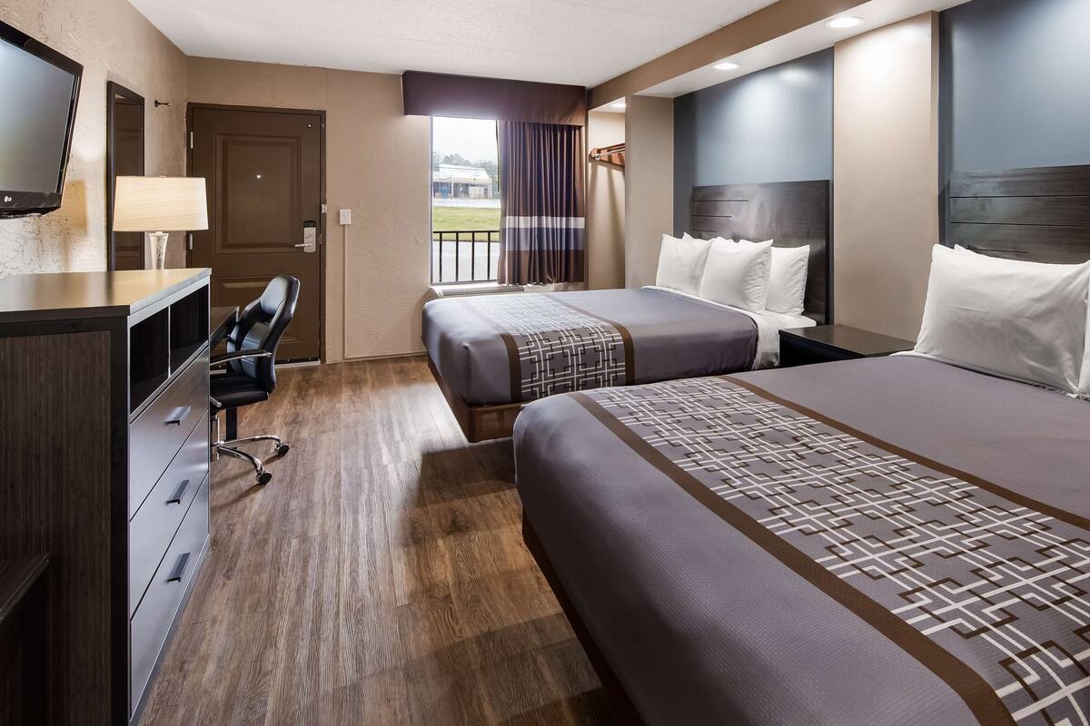 Habitación de hotel con dos camas, un escritorio con silla, televisión y vistas a un edificio.