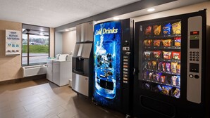 En selvbetjeningsautomat med et udvalg af snacks og kolde drikkevarer, en vanddispenser og et skilt, der angiver tilgængelige faciliteter som sæbe, blegemiddel og blødgøringsmiddel.