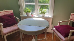 Una habitación con dos sillas blancas con cojines rojos, una mesa redonda pequeña con mantel azul claro y dos plantas en macetas en un alféizar.