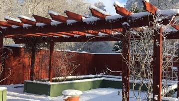 A snow-covered outdoor area with a wooden pergola structure, green planters, and a small tree.