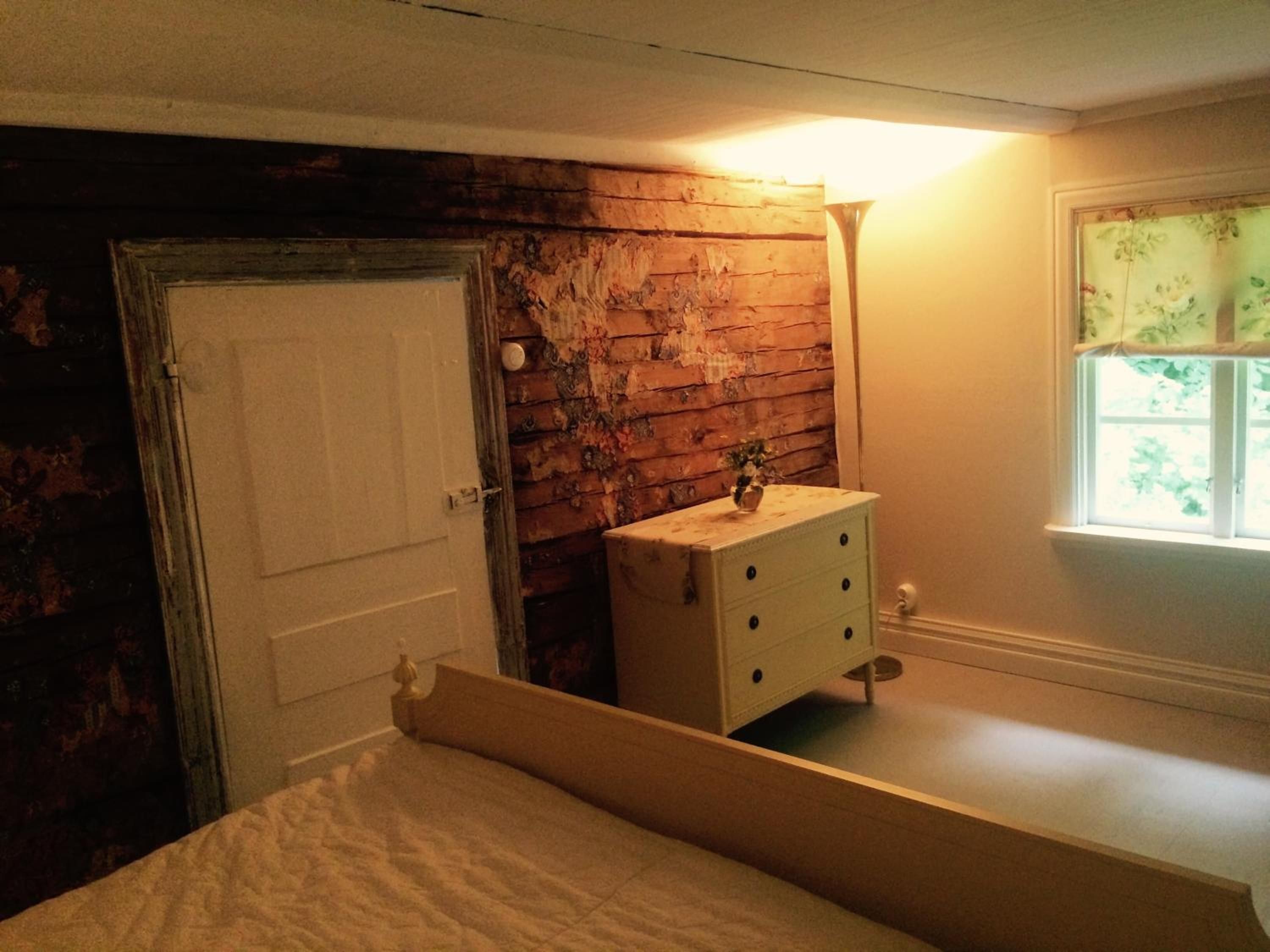 Un dormitorio con pared de madera, una puerta blanca, una mesita de noche con lámpara y una ventana con vistas.