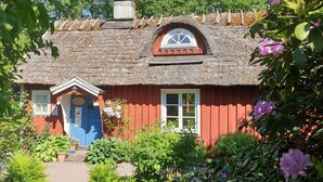 Una cabaña tradicional con techo de paja, fachada roja, puerta azul y un pequeño jardín con diversas plantas y flores.