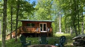 Eine Hütte mit einer Veranda, umgeben von einem Wald. Im Vordergrund befindet sich eine Feuerstelle.