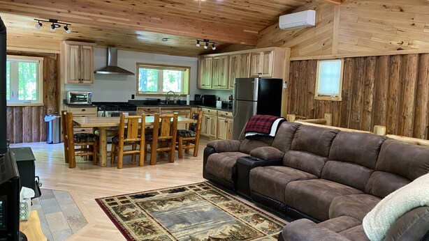 Ein gemütliches Blockhaus-Interieur mit einer Holzespeisegruppe, einem braunen Ledersofa und einer Küche mit Holzschränken und einem Kühlschrank.