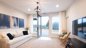 A modern living room with a white sofa, a flat-screen TV, and a large window offering a mountain view.
