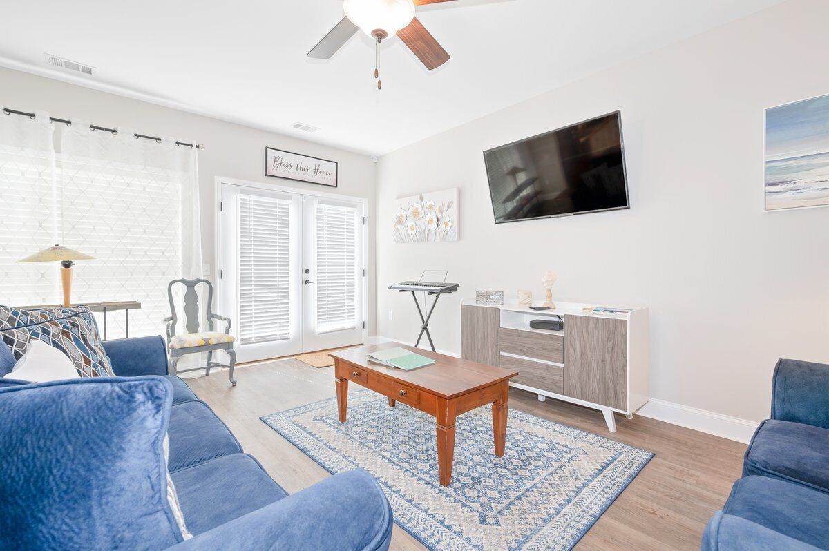 A living room with a blue sofa, a wooden coffee table, a TV mounted on the wall, and a ceiling fan.
