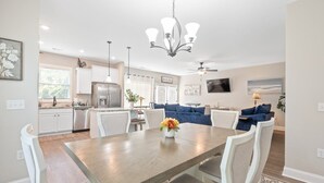 A modern kitchen with white cabinets, a stainless steel refrigerator, and a dining area with a wooden table and white chairs. - Gorgeous new home, one level & convenient location (Brunswick)