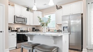 A modern kitchen with white cabinets, stainless steel appliances, a marble island, and two bar stools. - Gorgeous new home, one level & convenient location (Brunswick)