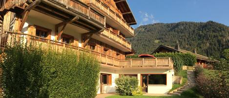 A wooden chalet with a balcony, a staircase leading to the entrance, and a mountain backdrop.
