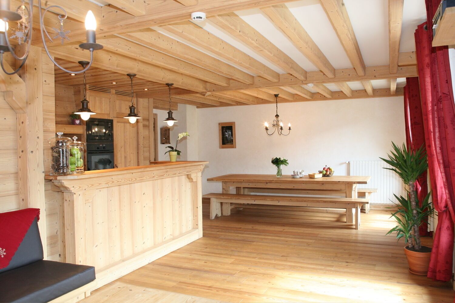 Un intérieur en bois comprenant un coin cuisine, des poutres apparentes au plafond, une table à manger avec des bancs et des plantes en pot.