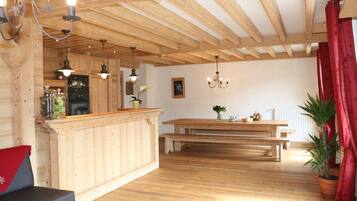 Un intérieur en bois comprenant un coin cuisine, des poutres apparentes au plafond, une table à manger avec des bancs et des plantes en pot.