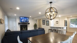 A modern living room with a dark-colored sectional sofa, a wooden dining table, white chairs, a TV mounted on the wall, and a chandelier. - Centrally Located DoCo Oasis (Fish Creek)