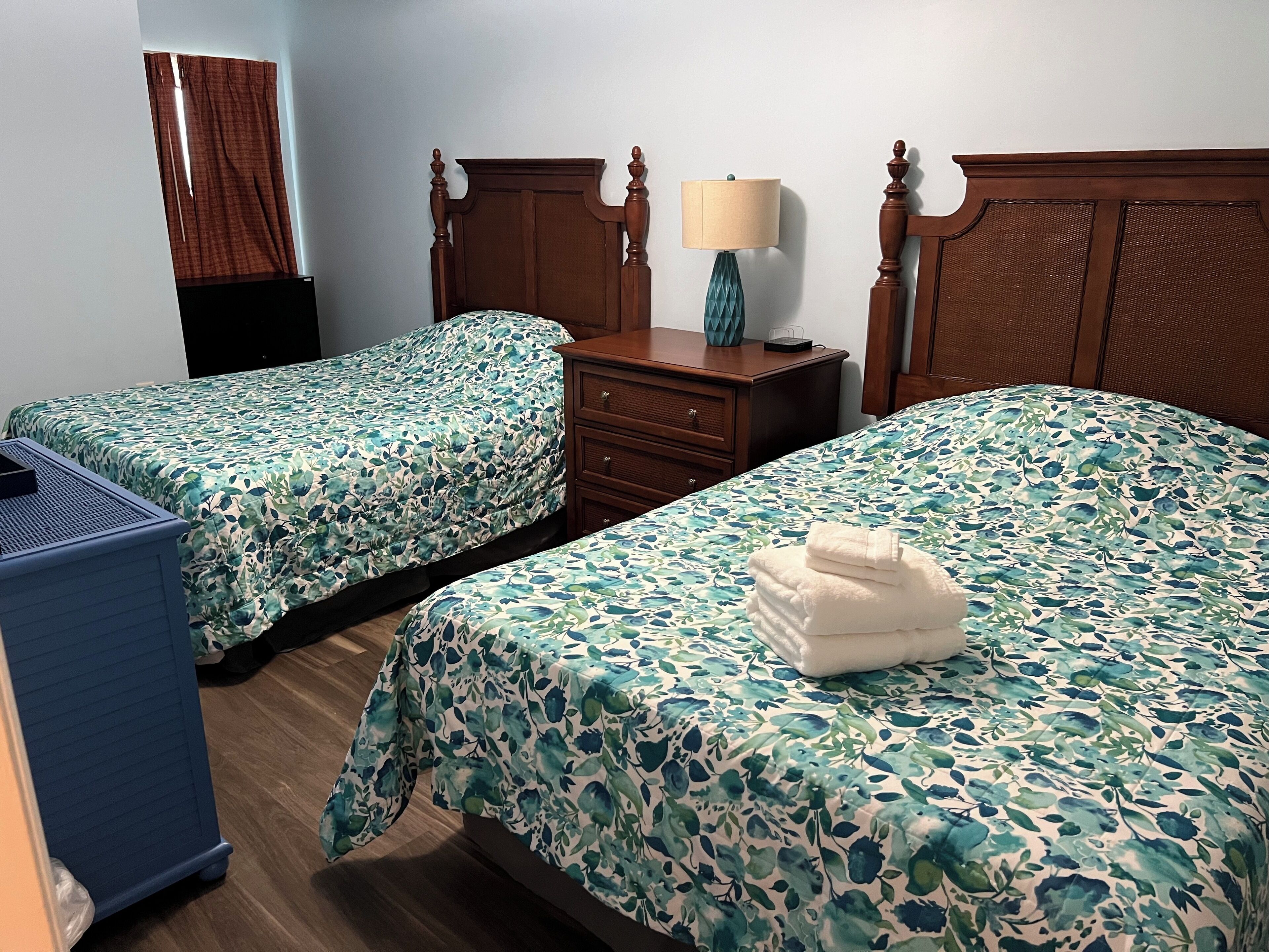 A hotel room with two single beds, a wooden dresser, a lamp, and a blue storage unit.
