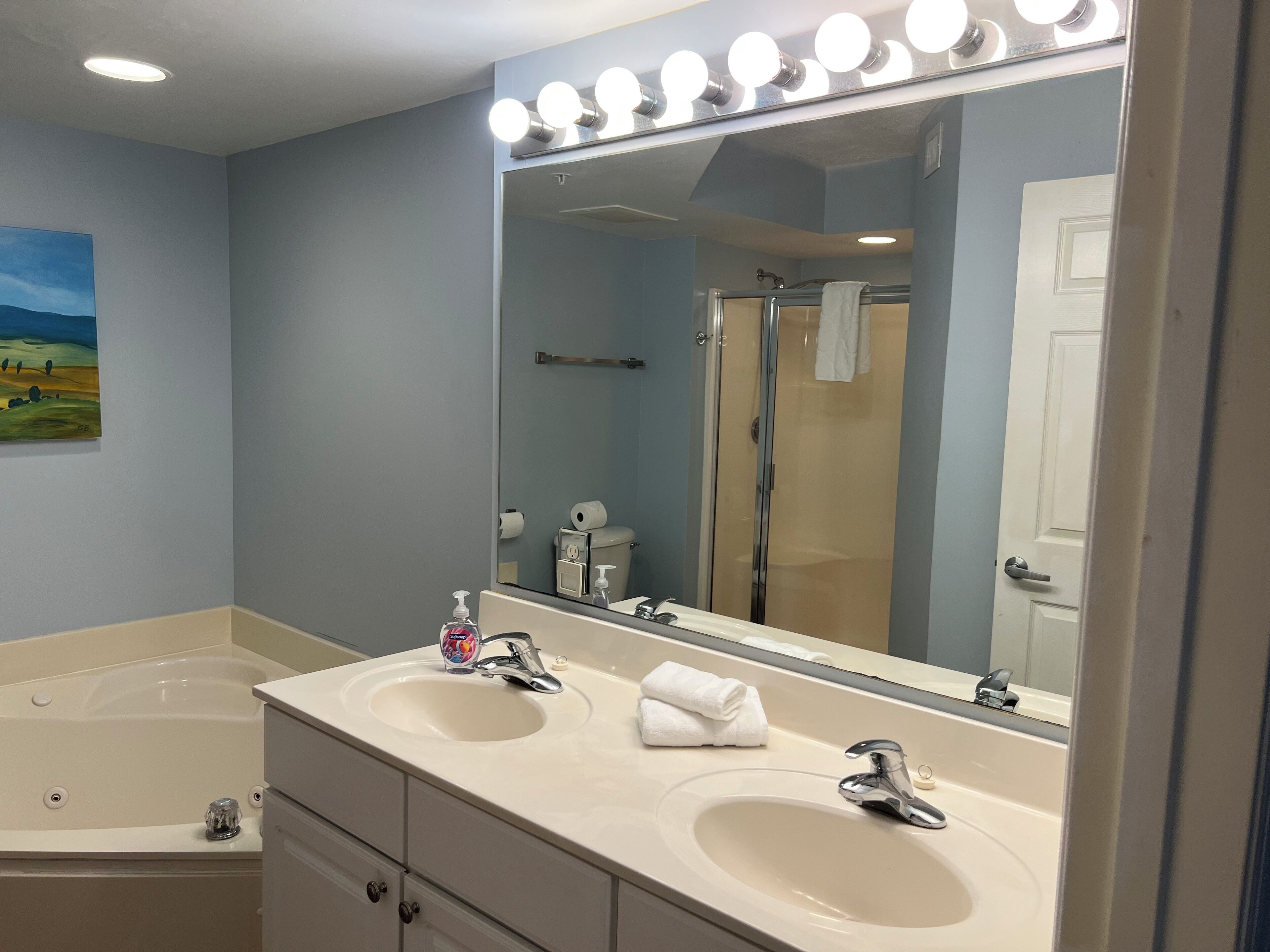 A bathroom with two sinks, a large mirror with lights, a shower, and a painting on the wall.