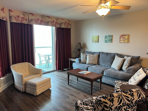 A living room with a grey sofa, a white armchair, a patterned armchair, a wooden coffee table, and a ceiling fan.