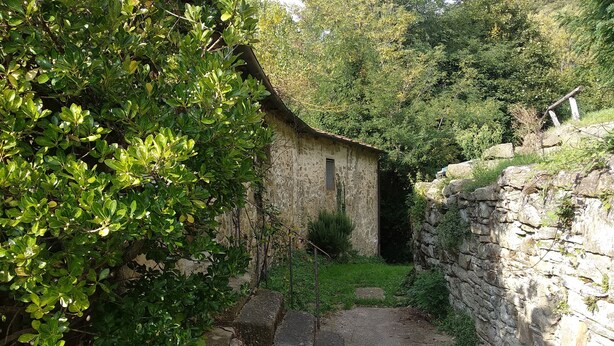 Ein rustikales Steinhaus mit schmalem Eingang, umgeben von üppiger Vegetation und einer Steinmauer.