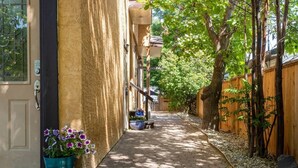 Une ruelle étroite avec un chemin pavé, une clôture en bois et un arbre qui offre de l’ombre.