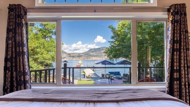 Une chambre avec une grande fenêtre donnant sur une terrasse au bord du lac, équipée de meubles d’extérieur et de parasols.